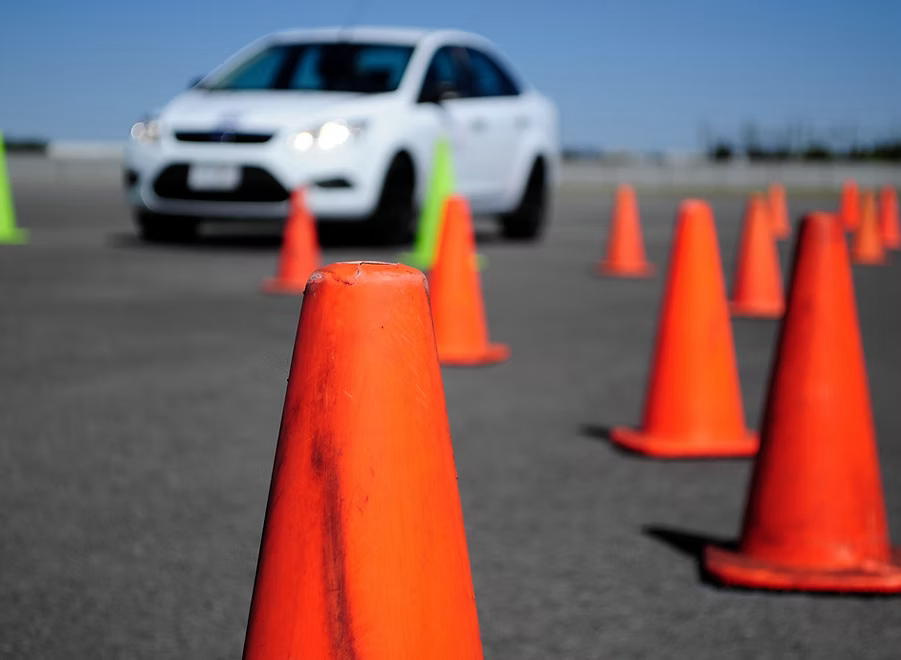 A series of bright orange traffic cones are arranged for a road test while a white car is visible in the distance